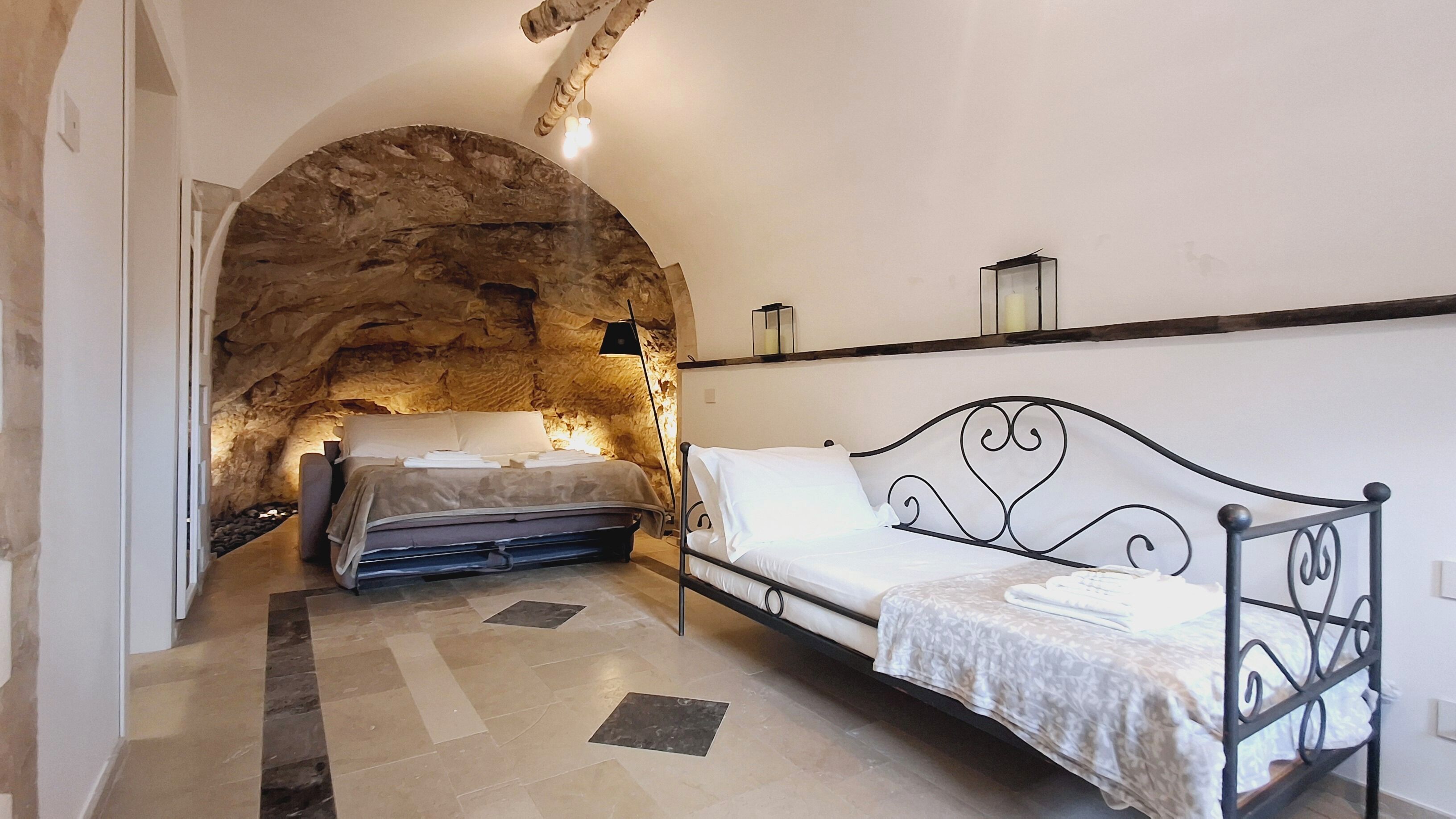 Un dormitorio con un arco de piedra, una cama con estructura metálica, una mesita de noche y un estante en la pared con una vela.
