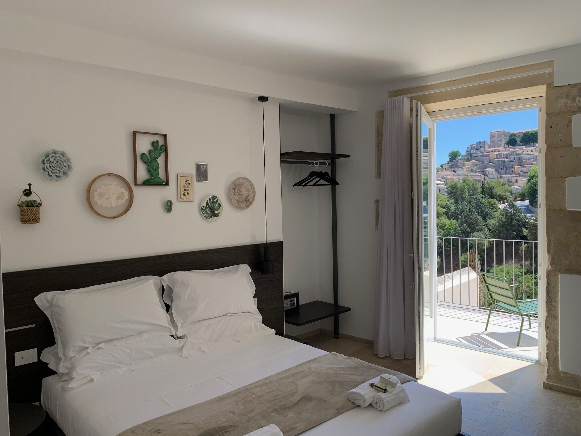 A bedroom with a bed, a bedside table, a hanging coat rack, and a balcony with a view of buildings.
