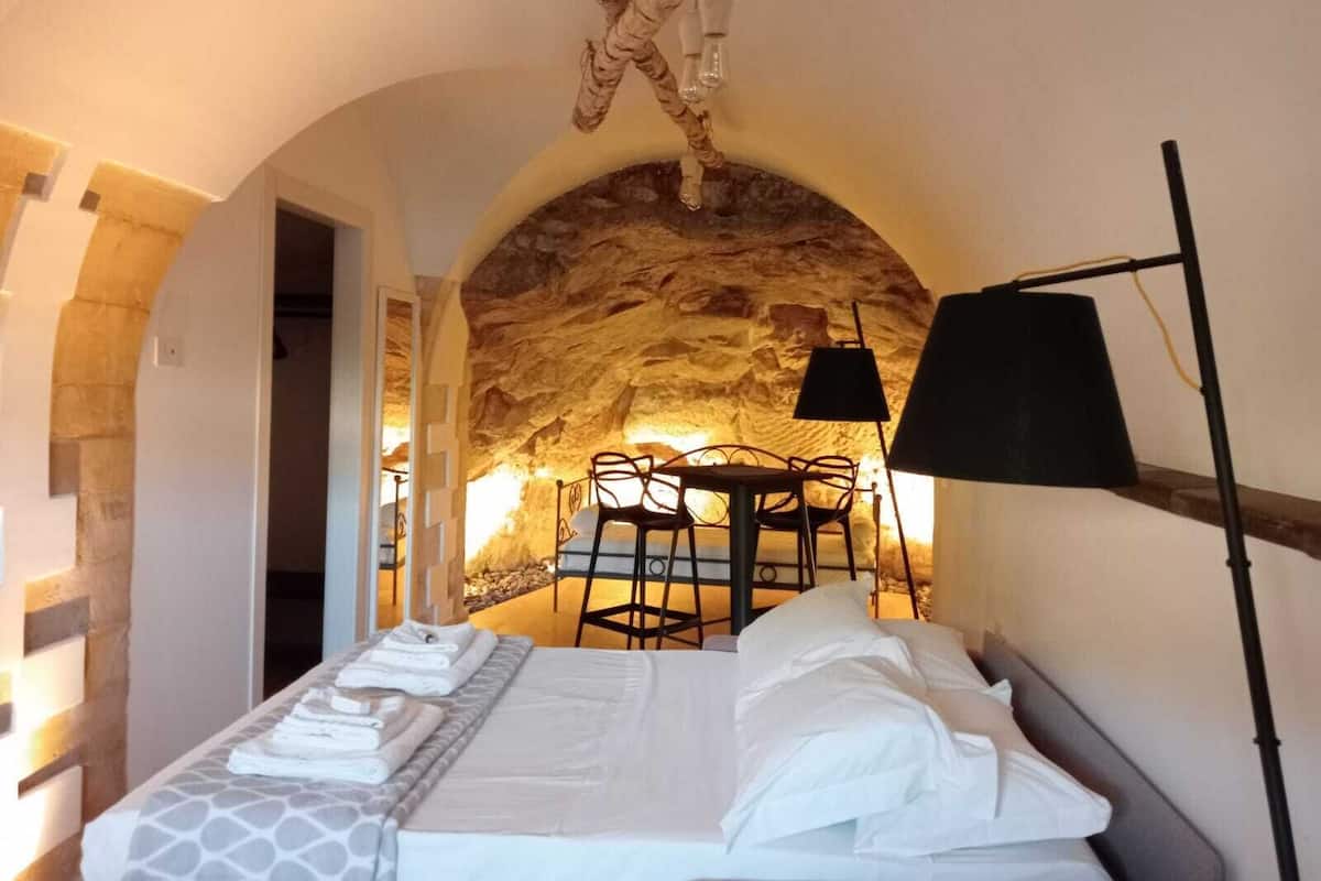 Ein Schlafzimmer mit einem Steinbogen, einem Bett mit weißen Leinen, einer schwarzen Lampe und einem Tisch mit Stühlen.