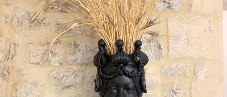 A black vase with wheat stalks on a black square base against a stone wall.