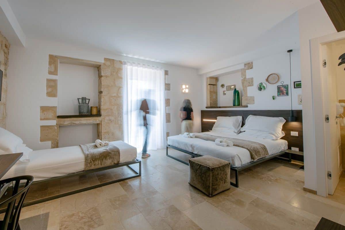 Ein modernes Hotelzimmer mit zwei Betten, einem kleinen Tisch und einem Stuhl. Es gibt ein großes Fenster mit Vorhängen, einen Spiegel an der Wand und eine Holzablage mit Dekoration.
