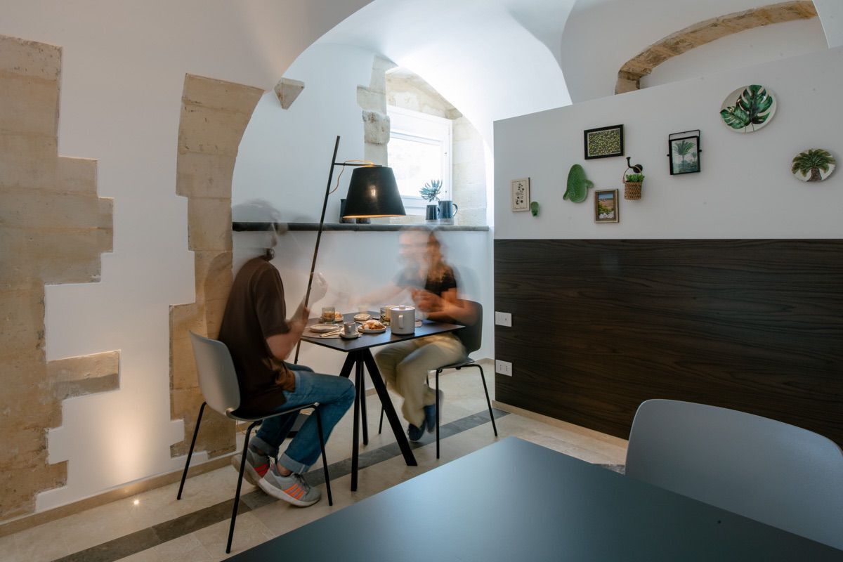 Comedor con una mesa, sillas y una persona sentada. Se observa una ventana con luz natural y una pared decorada con cuadros y plantas.