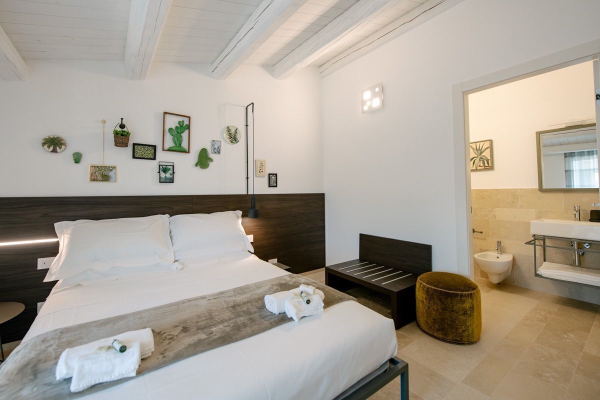 Una habitación de hotel moderna con una cama grande, una mesita de noche, un taburete pequeño, un banco de madera y un baño con lavabo y bidé.