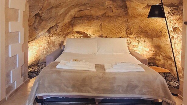 Un dormitorio con techo abovedado de piedra, una cama con colcha color beige, una lámpara de pie negra y una mesita de noche con lámpara.
