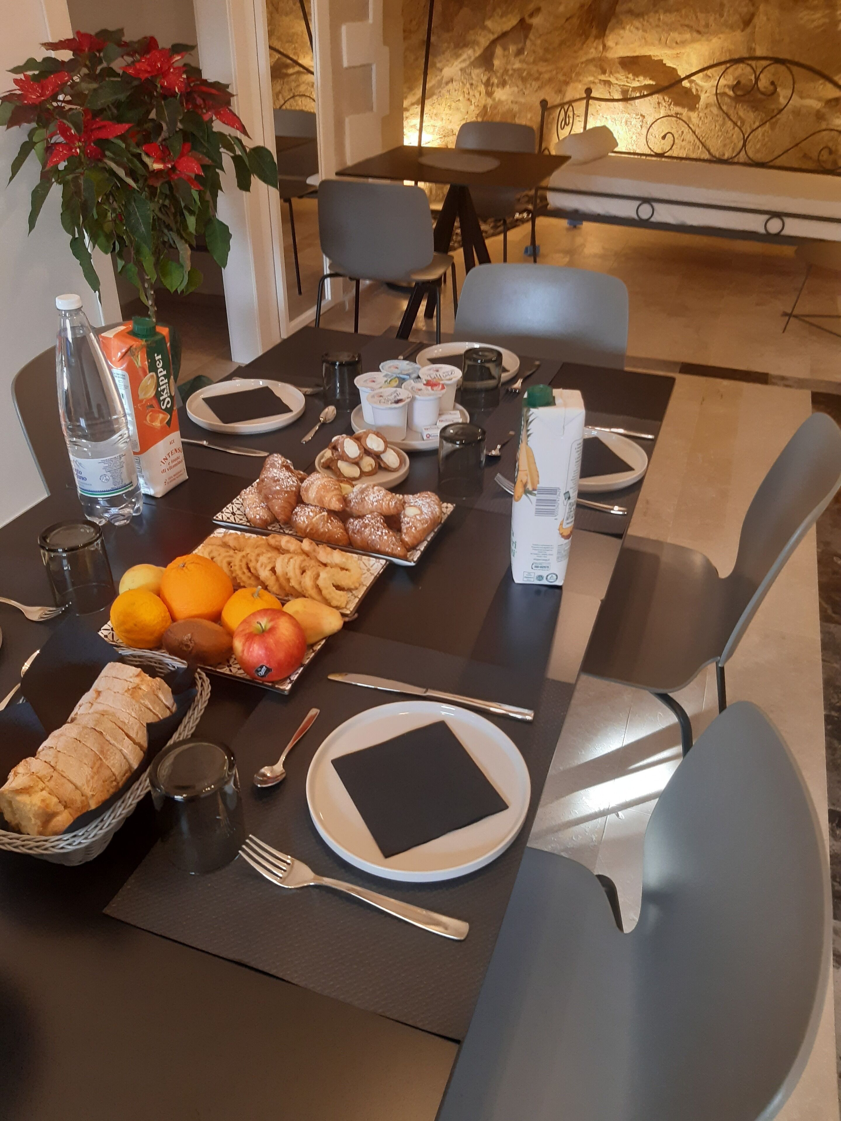 Una mesa de desayuno preparada con croissants, bollería, frutas y un cartón de jugo.