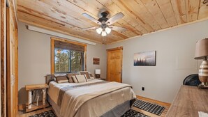 A bedroom with a bed, wooden headboard, a ceiling fan, a window with blinds, a wooden door, a painting on the wall, and a bedside table with a lamp.