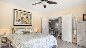 Une chambre à coucher avec un lit, deux tables de chevet avec des lampes, un ventilateur de plafond, une porte donnant sur une autre pièce et un tableau encadré au mur.
