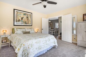A bedroom with a bed, two bedside tables with lamps, a ceiling fan, a door leading to another room, and a framed picture on the wall.