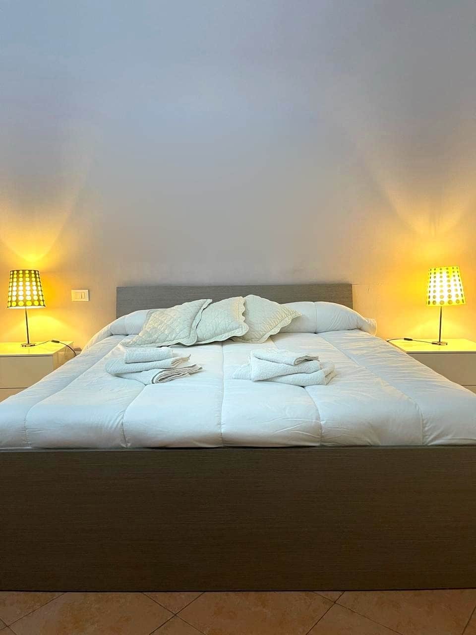 Ein ordentlich bezogenes Bett mit Kissen und Decke, zwei Nachttischlampen und eine schlichte Wand.