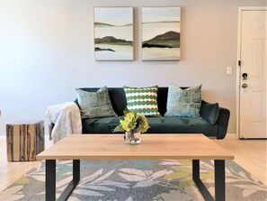 A living room with a wooden coffee table, a green sofa with patterned cushions, and two landscape paintings on the wall.