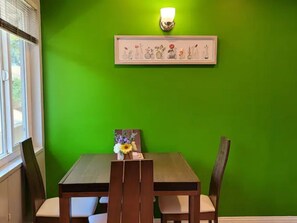 A dining area with a green wall, a framed artwork above the table, and a vase with flowers on the table.