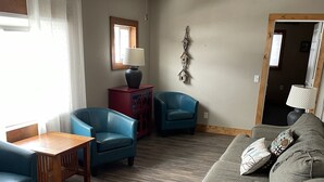 Un salon avec un ventilateur de plafond, une table en bois, deux chaises bleues, un canapé gris agrémenté de coussins à motifs et un buffet rouge.