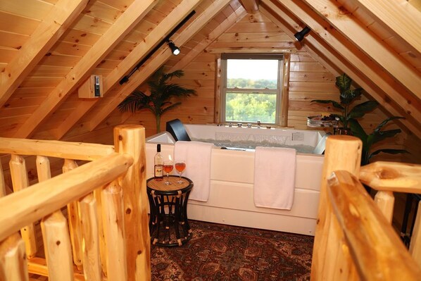 A cozy attic bedroom with a wooden ceiling, a large window, a bathtub, and a small table with drinks.