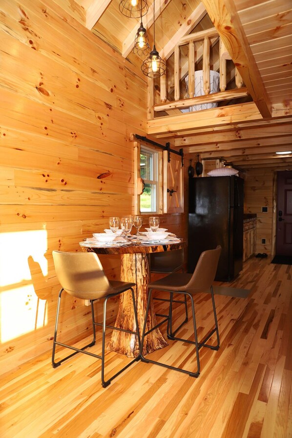 Interior de una cabaña de madera con una mesa de comedor preparada para cuatro personas, dos sillas, un refrigerador negro y una ventana con vistas a la vegetación.