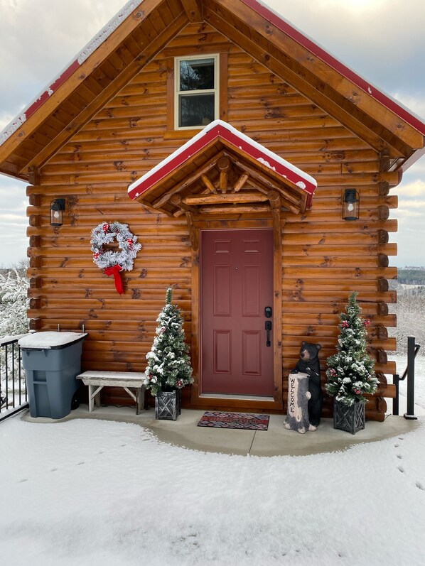 A log cabin with a red door, a wreath, two Christmas trees, a bench, and a trash bin.