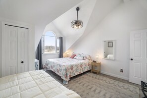Une chambre avec un plafond en pente, un lit recouvert d’une couvre-lit à motifs floraux, une table de chevet avec une lampe et une fenêtre donnant sur un beau paysage.