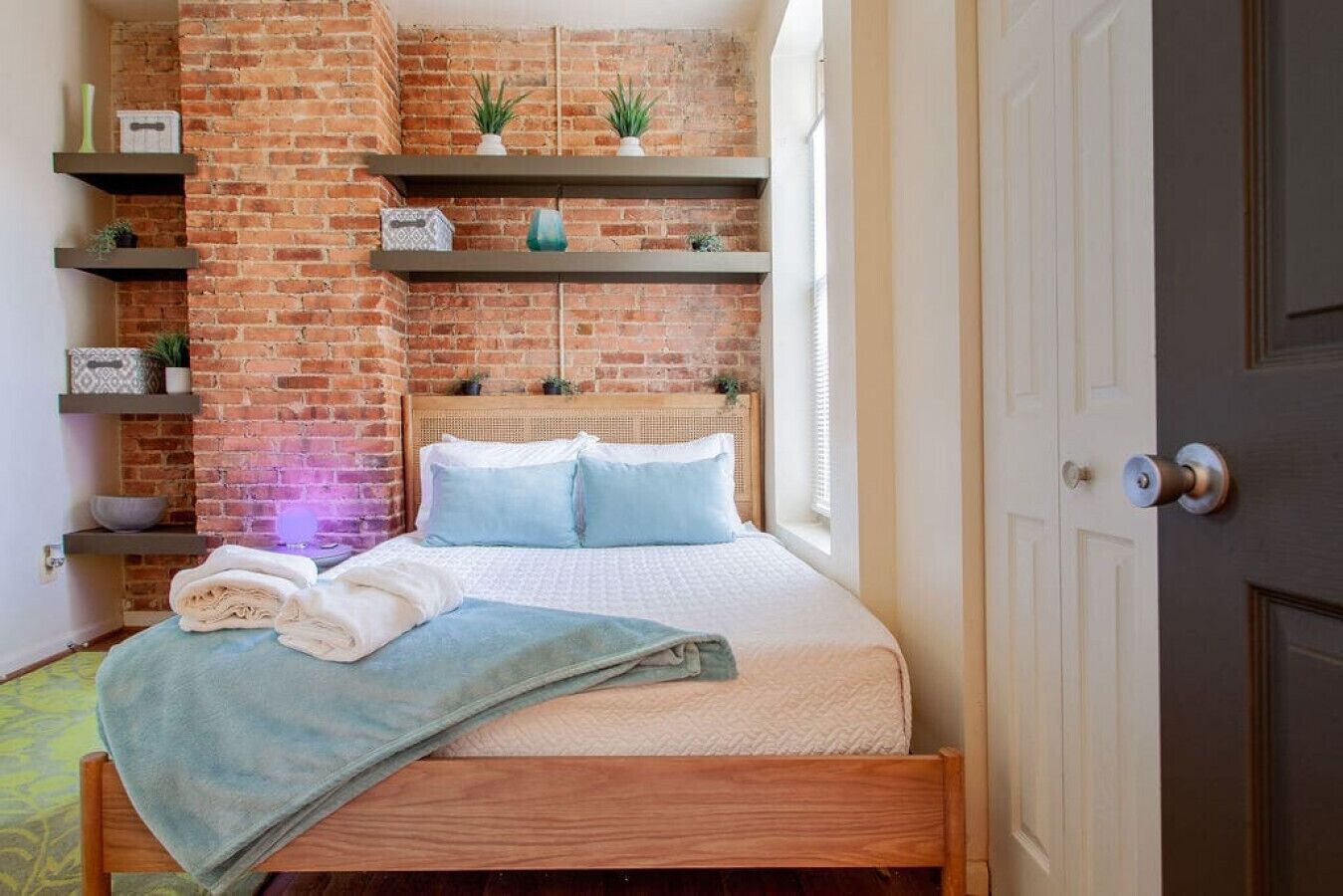 A bedroom with a brick wall, a bed with blue bedding, and shelves with plants and decorative items.