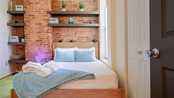 A bedroom with a brick wall, a bed with blue bedding, and shelves with plants and decorative items.