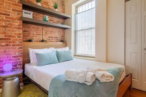 A bedroom with a brick wall, a bed with a white and blue bedspread, a bedside table with a lamp, and a window with blinds. - Colorful & Quirky 2-BR Loft w/Private Deck | MICA, JH & UMMC (Baltimore)