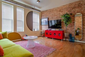 A living room with a brick wall, a red cabinet, a TV, a round table, and a green sofa. - Colorful & Quirky 2-BR Loft w/Private Deck | MICA, JH & UMMC (Baltimore)
