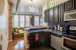 A compact kitchen with dark cabinets, a stainless steel sink, and a built-in oven. - Colorful & Quirky 2-BR Loft w/Private Deck | MICA, JH & UMMC (Baltimore)