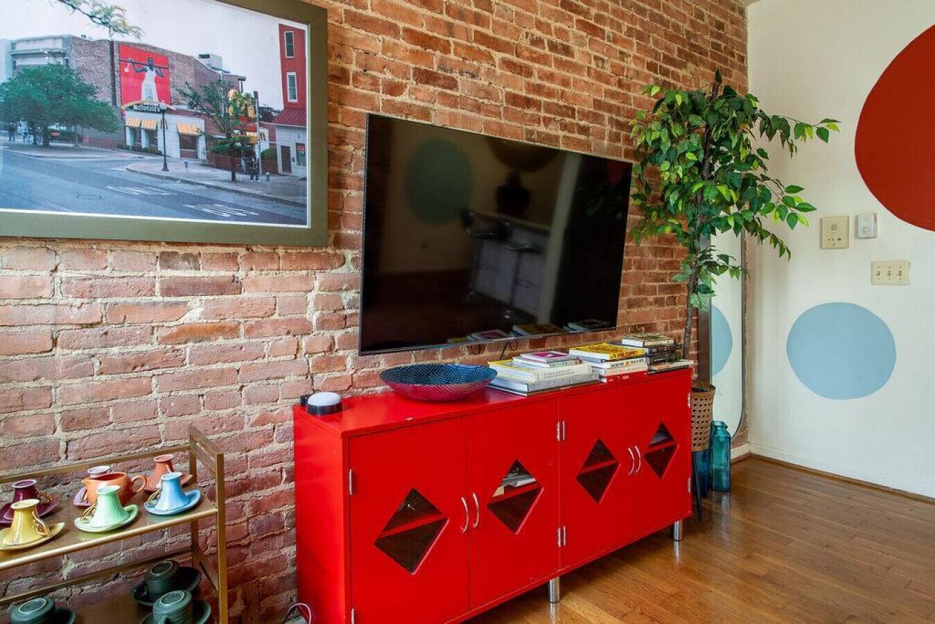 Una sala de estar con pared de ladrillo, un mueble rojo con libros, una televisión y una planta.