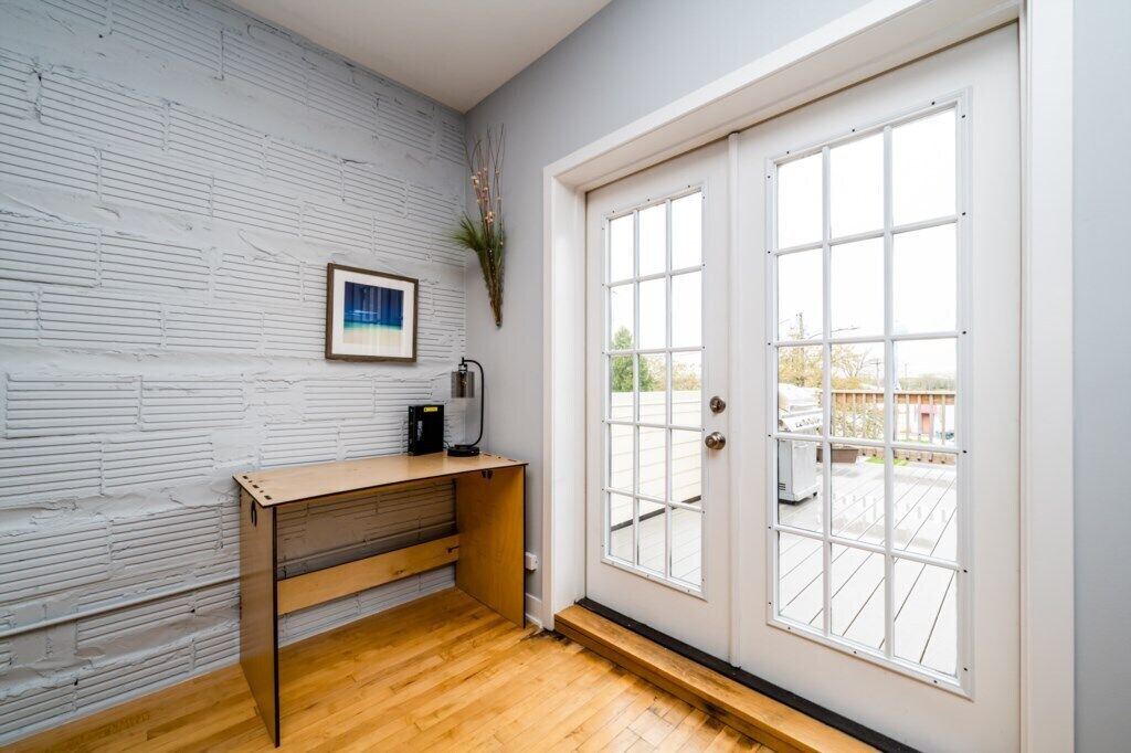A room with a white brick wall, a wooden desk, a framed picture, and a large window with a view of the outside.