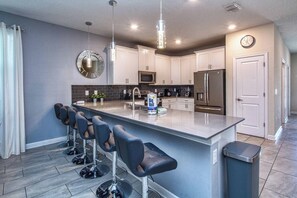 A modern kitchen with a central island, bar stools, a microwave, a refrigerator, and a clock on the wall.