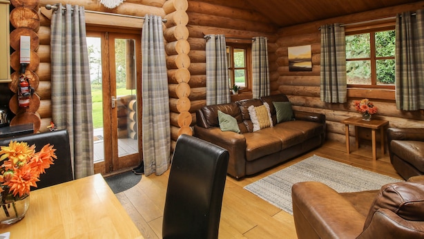 A log cabin interior with a wooden floor, a leather sofa, plaid curtains, and a wooden table with a vase of flowers. - Brynallt Country Park (Ellesmere)