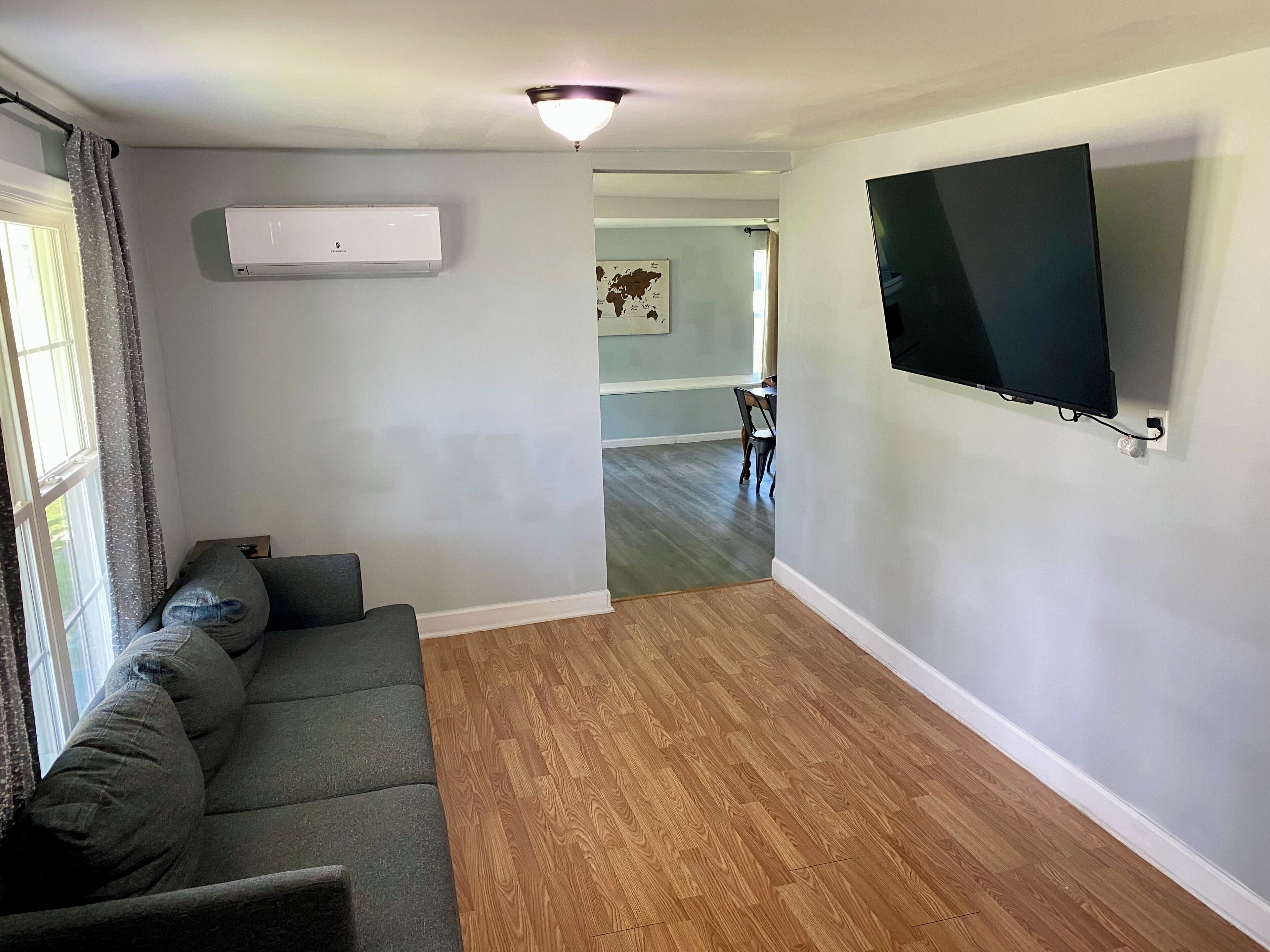 A living room with a gray sofa, wooden flooring, a mounted TV, and an air conditioning unit.
