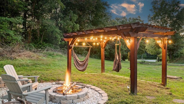 Een houten pergola met lichtjes, een vuurkorf en een hangstoel in een gazon.