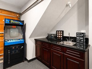 Une petite cuisine équipée comprenant un distributeur de jeux d’arcade rétro, un évier et un panneau affichant un message humoristique.