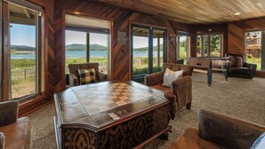 A spacious room with a large wooden table, comfortable seating, and large windows offering a view of a lake and hills. - Grand Lakefront Escape - Lakefront Cabin | Hot Tub, Pool Table (Big Bear Lake)