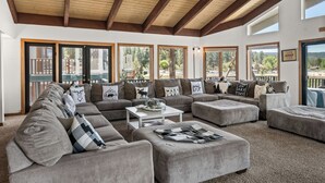 A spacious living area with multiple sofas and ottomans, large windows with a view of a pool and trees, and a ceiling fan. - Grand Lakefront Escape - Lakefront Cabin | Hot Tub, Pool Table (Big Bear Lake)