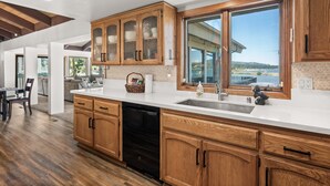 A modern kitchen with wooden cabinets, a dishwasher, and a large window overlooking a scenic view. - Grand Lakefront Escape - Lakefront Cabin | Hot Tub, Pool Table (Big Bear Lake)