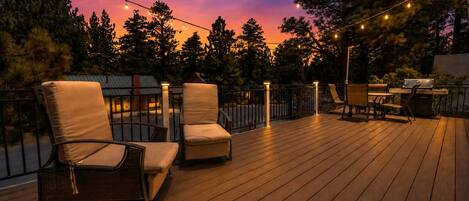 An outdoor deck with wooden flooring, two cushioned chairs, a grill, and string lights against a twilight sky.