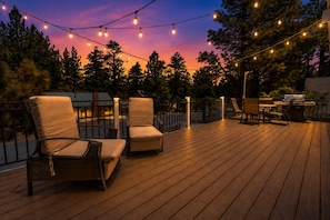 An outdoor deck with wooden flooring, two cushioned chairs, a grill, and string lights against a twilight sky.