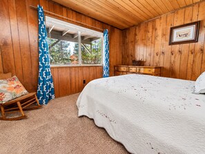 Un dormitorio con una cama grande, paredes de madera, un ventanal con cortinas, un aparador y una mecedora.