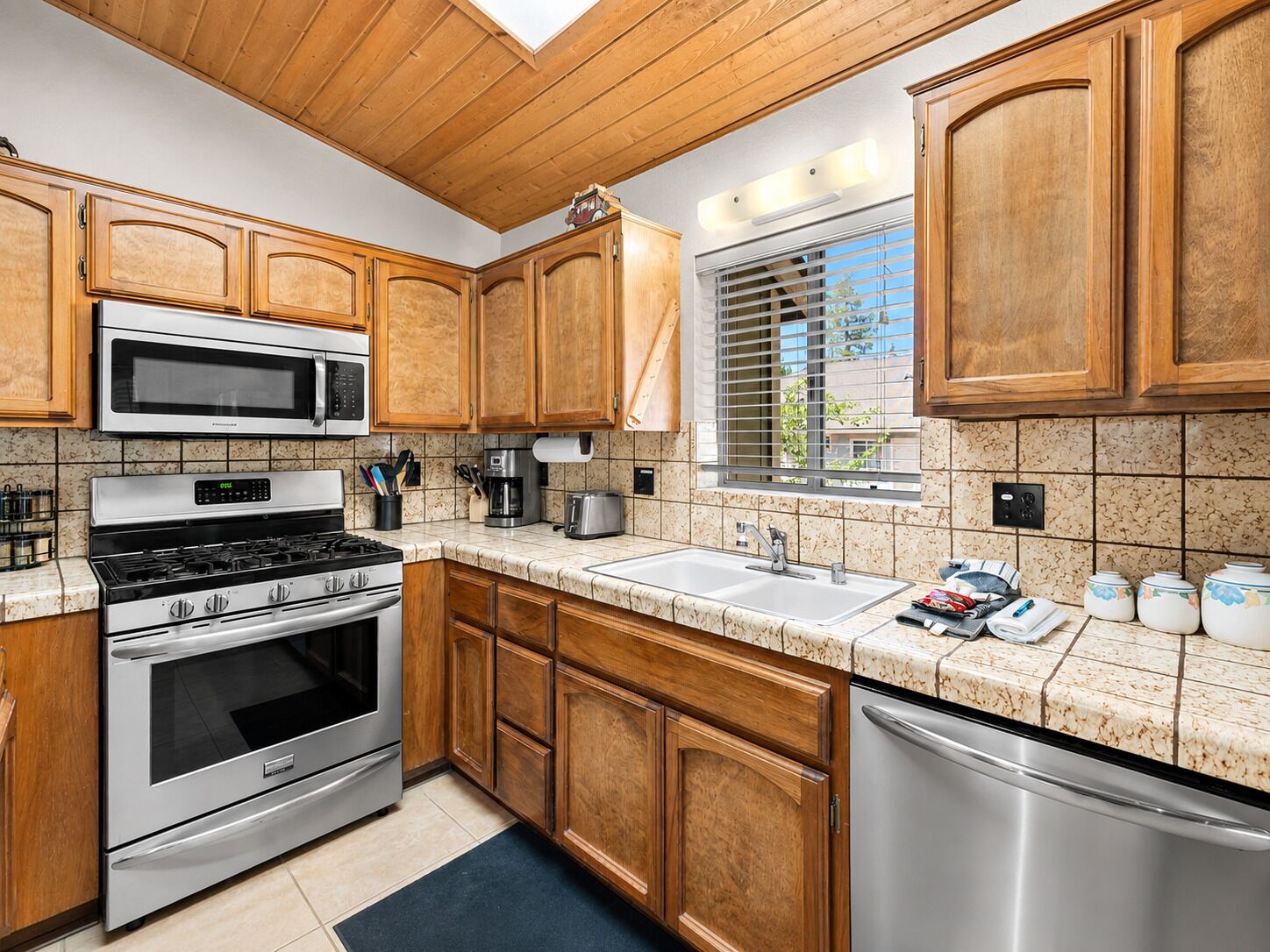 A kitchen with wooden cabinets, stainless steel appliances, a window with blinds, and a tiled backsplash.