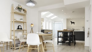 A modern kitchen with a wooden dining table, white chairs, and a black bar stool. There is a ladder leaning against the wall and a shelf with decorative items.