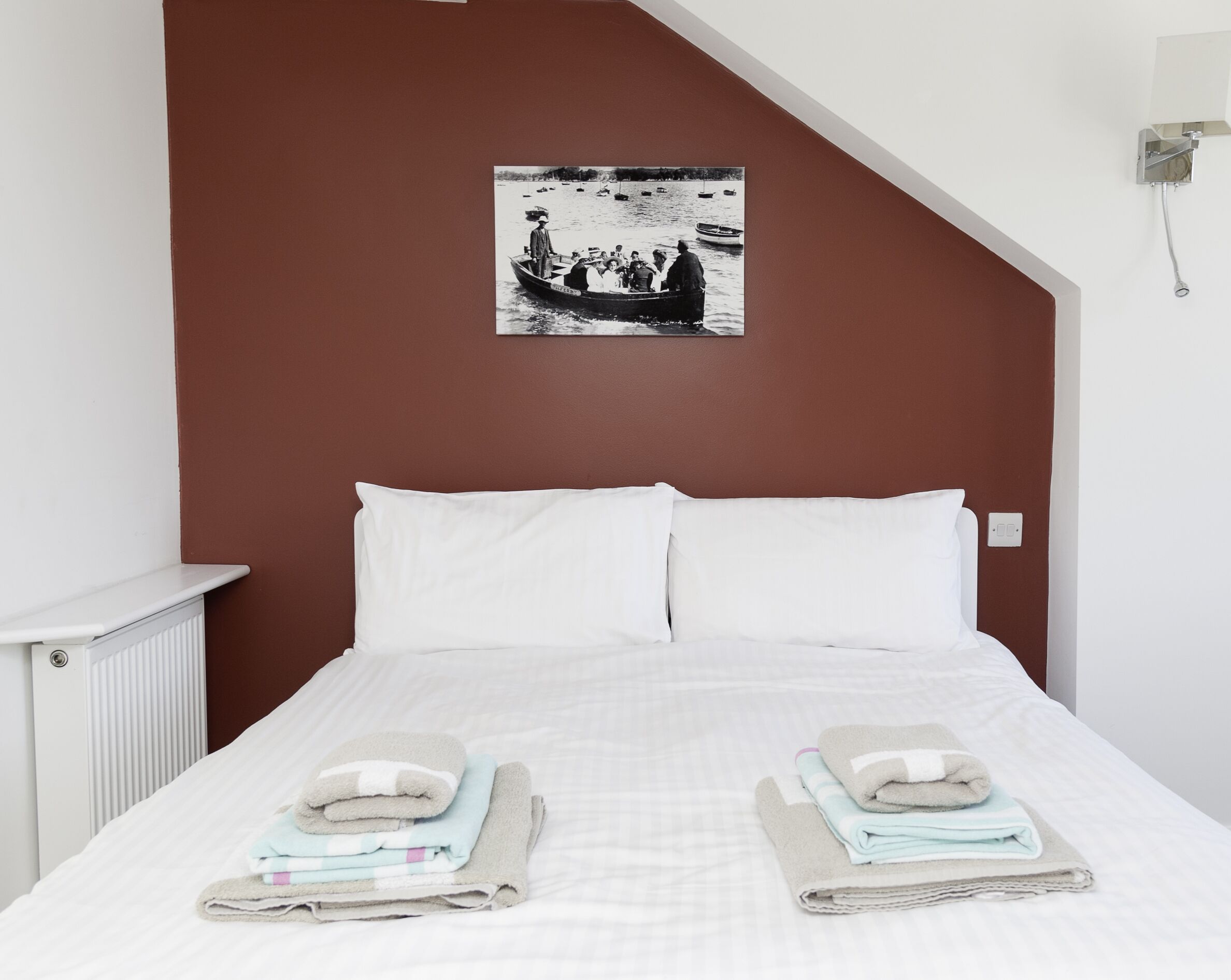 Un dormitorio con una cama, ropa de cama blanca, dos toallas dobladas, una mesita de noche blanca, un radiador y una fotografía blanco y negro en la pared.