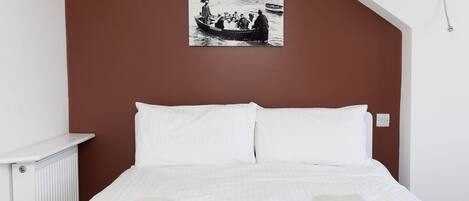 Un dormitorio con una cama, ropa de cama blanca, dos toallas dobladas, una mesita de noche blanca, un radiador y una fotografía blanco y negro en la pared.