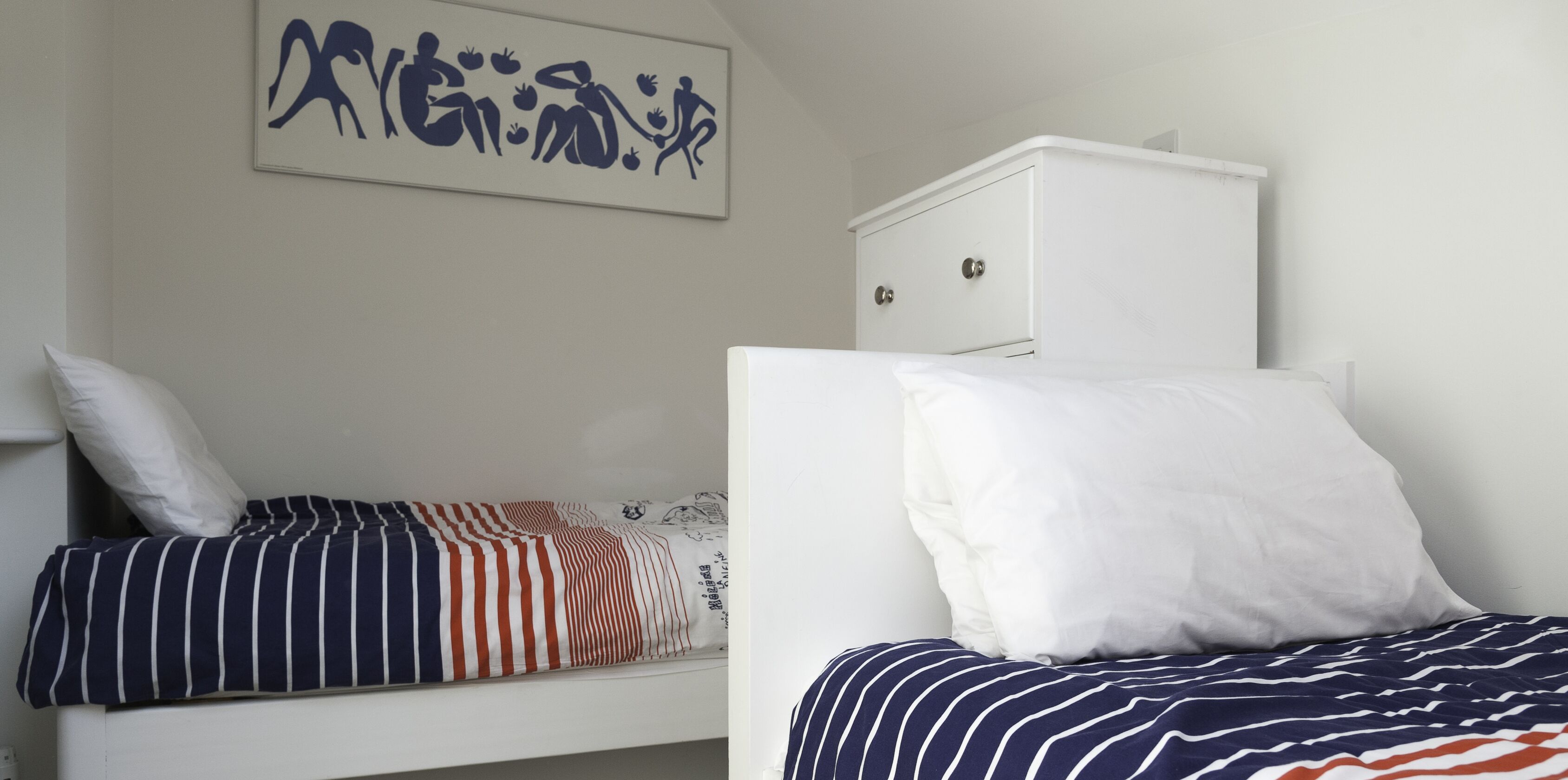 Deux lits simples avec du linge de lit à rayures, une armoire blanche et un mur orné d’une œuvre d’art abstraite bleue.
