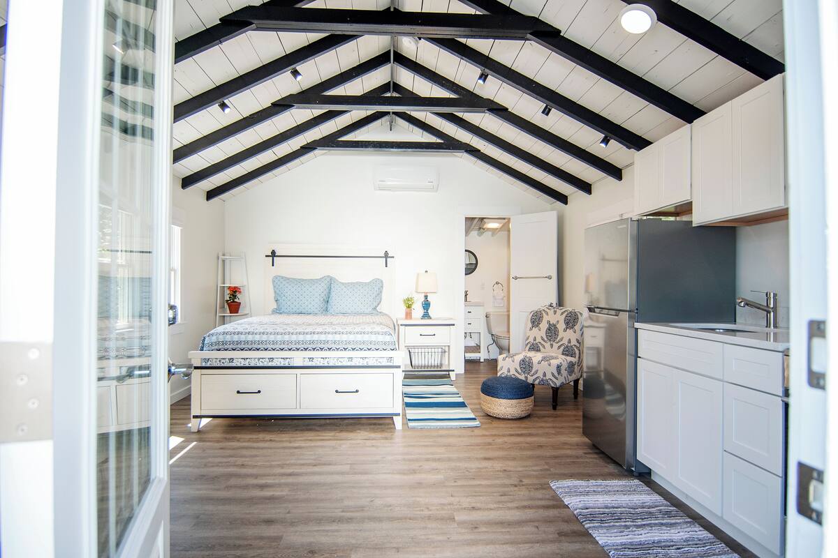 A bedroom with a large bed, bedside tables, a chair, and a kitchenette with a refrigerator.