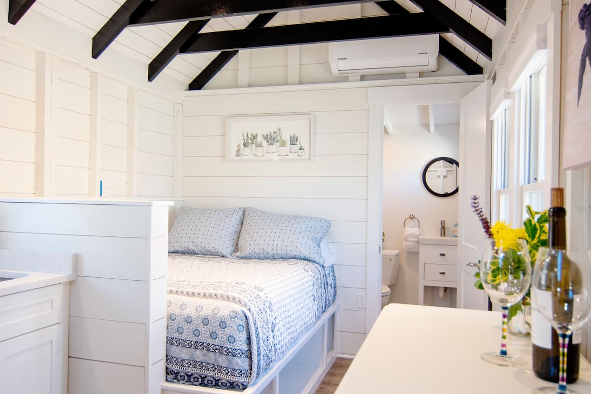 A bedroom with a bed, a bedside table with a wine bottle and glasses, a small bedside table with a vase of flowers, a wall-mounted clock, and a framed picture of plants.