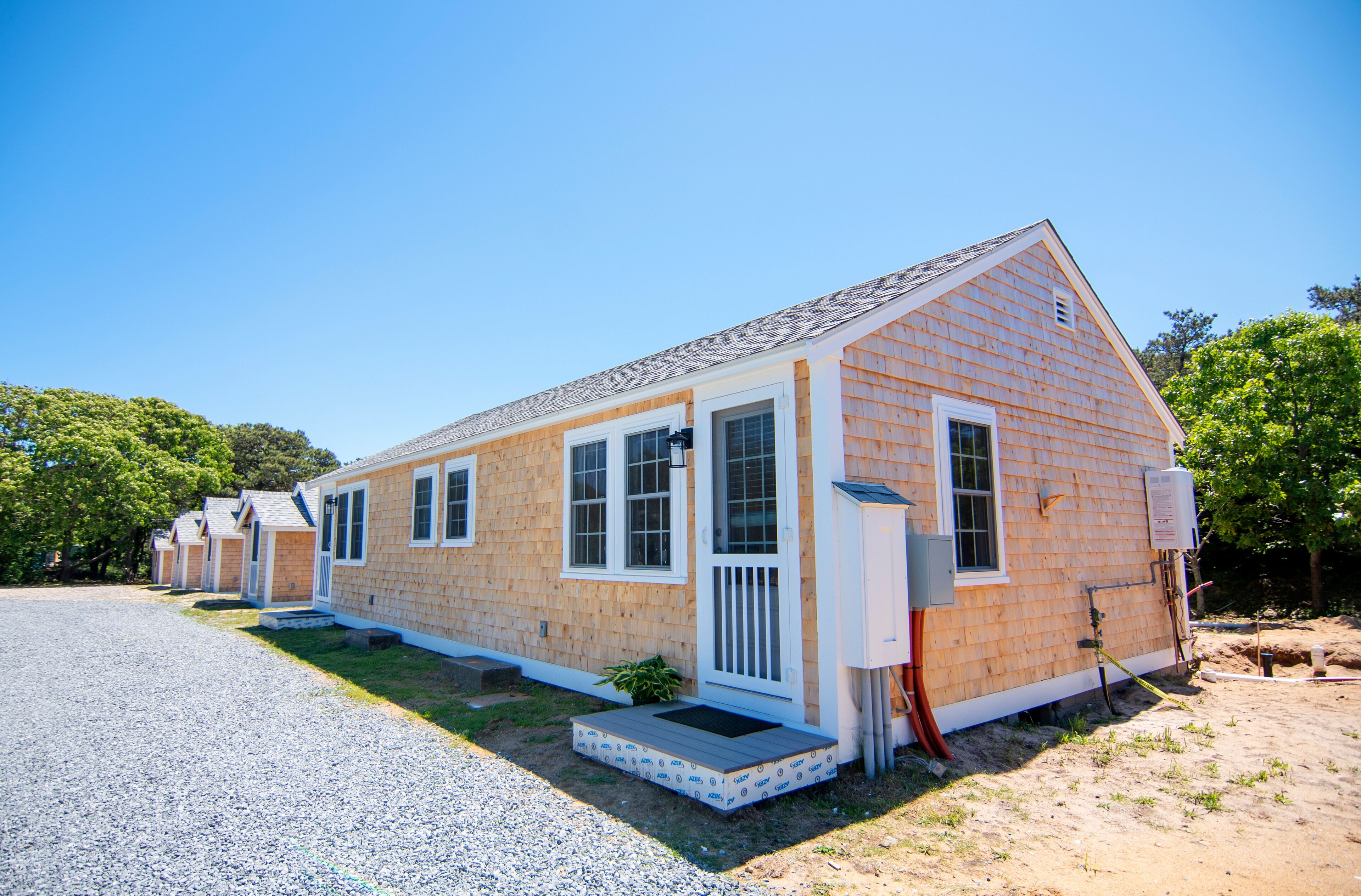 A row of small, single-story wooden cabins with a gravel driveway and a clear blue sky.