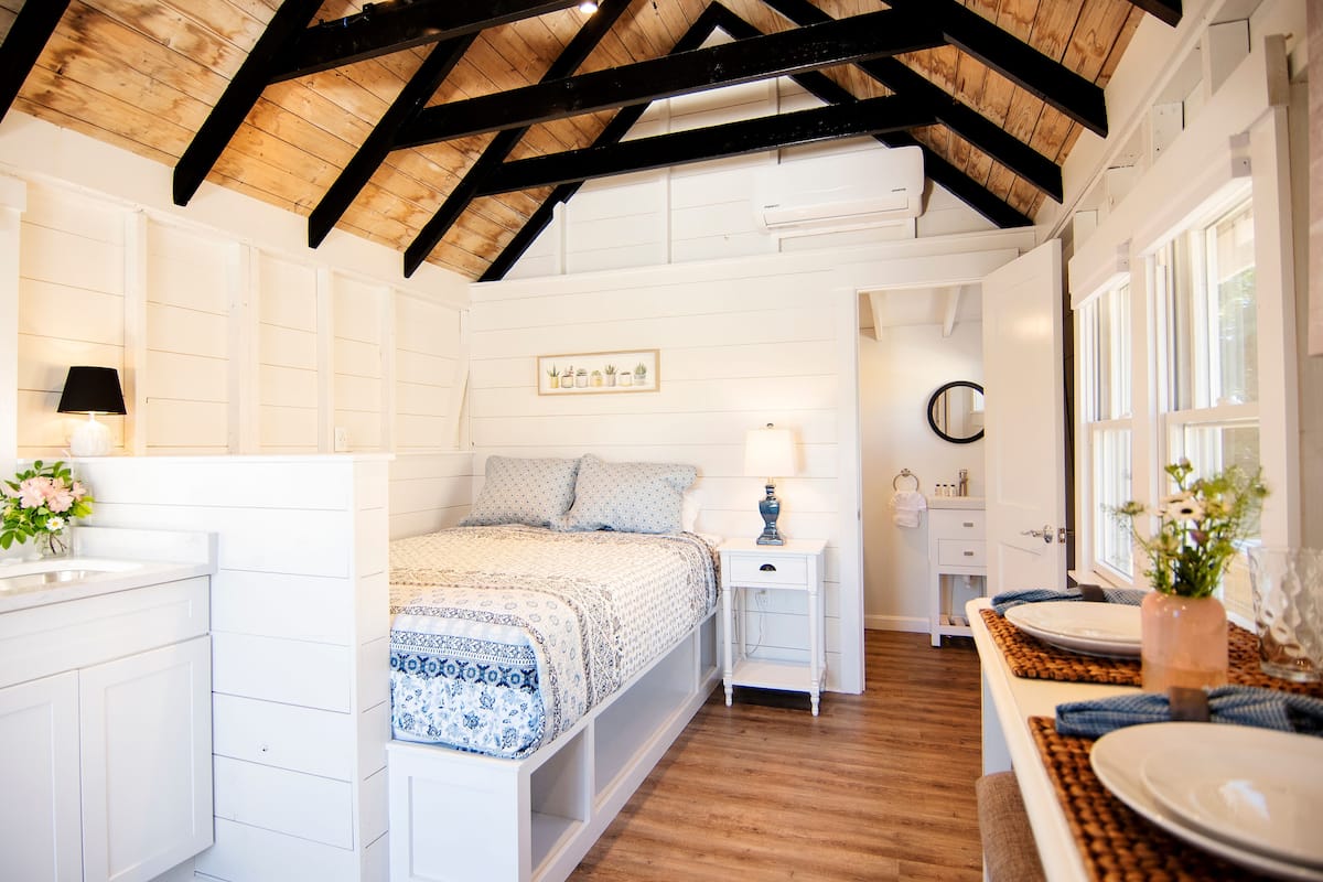 A bedroom with a wooden ceiling, a bed with a patterned bedspread, a bedside table with a lamp, and a window with a view of the outside.