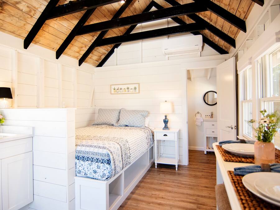 A bedroom with a wooden ceiling, a bed with a patterned bedspread, a bedside table with a lamp, and a window with a view of the outside.