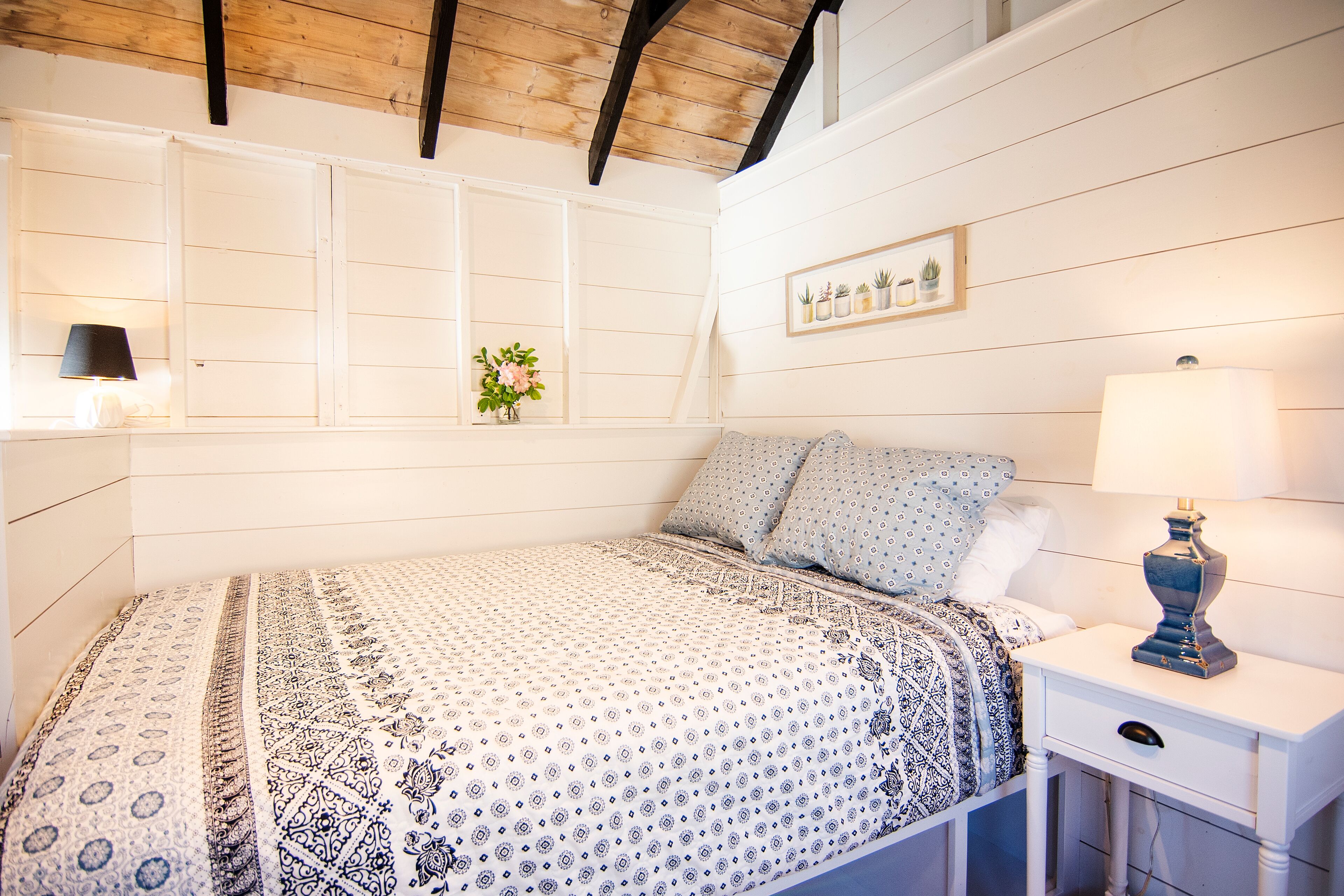 A bedroom with a patterned bedspread, a bedside table with a lamp, and a wall-mounted shelf with potted plants.