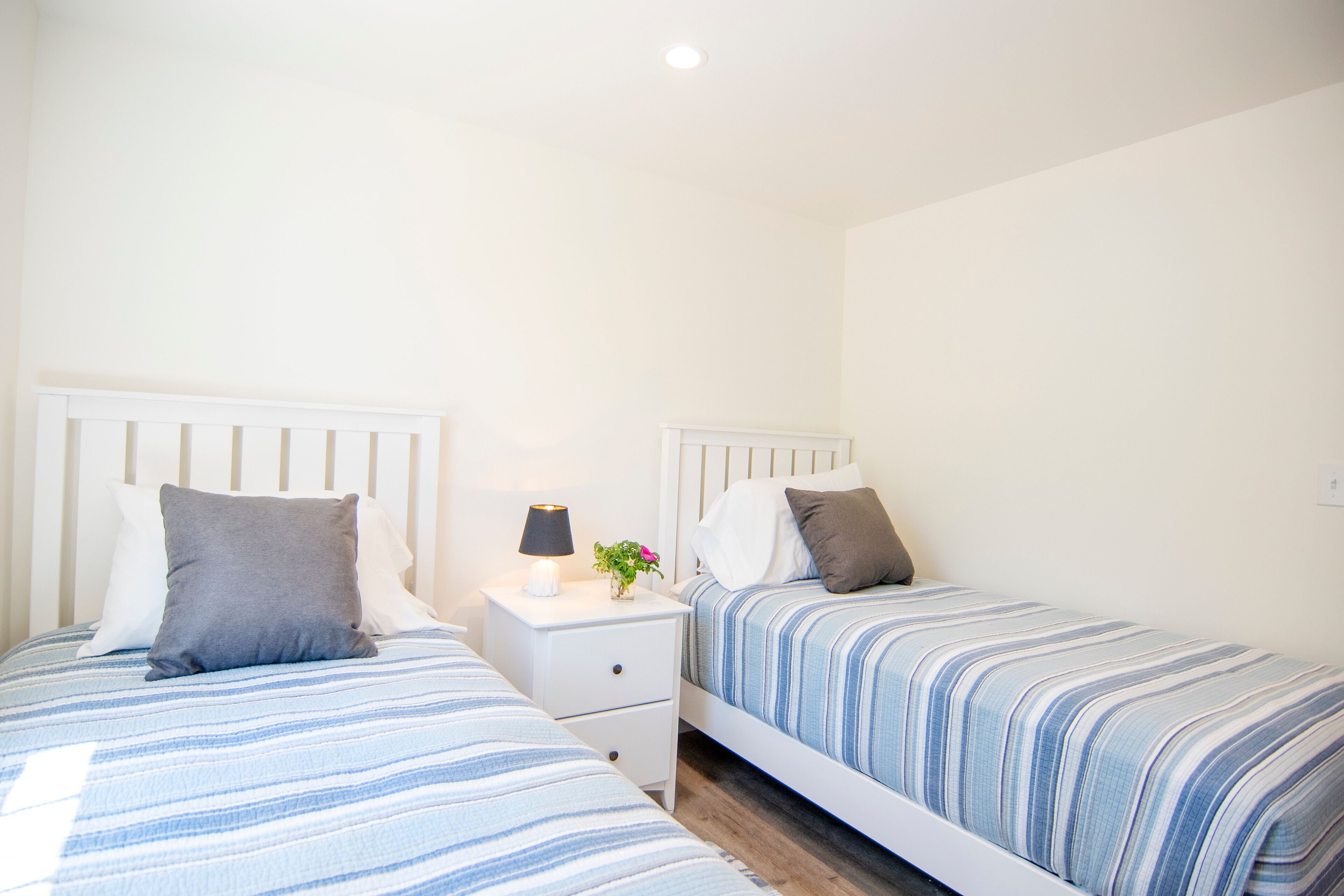 A twin bedroom with two beds, each with striped bedding, a bedside table with a lamp, and a small vase with flowers.